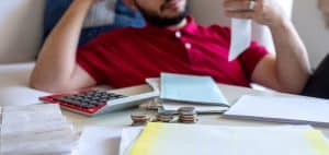 A man in a red shirt sits at a table, looking stressed while reviewing bills, receipts, and financial documents.